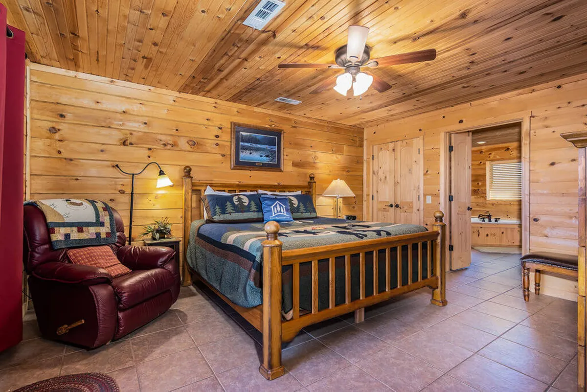 A cozy bedroom featuring a wooden bed frame, a recliner chair positioned for reading or relaxing, a decorative lamp on a nightstand, and framed nature-themed artwork on the wall. The space is bathed in warm light, with soft bedding that looks fresh and inviting.