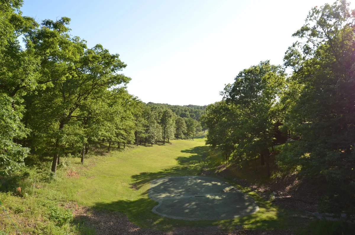 A lush green open field bordered by mature trees and a golf course area stretches into the distance, offering ample room for outdoor activities. The inviting, well-maintained lawn is surrounded by natural beauty, showcasing wide open spaces ideal for walking, playing, or relaxing with pets.