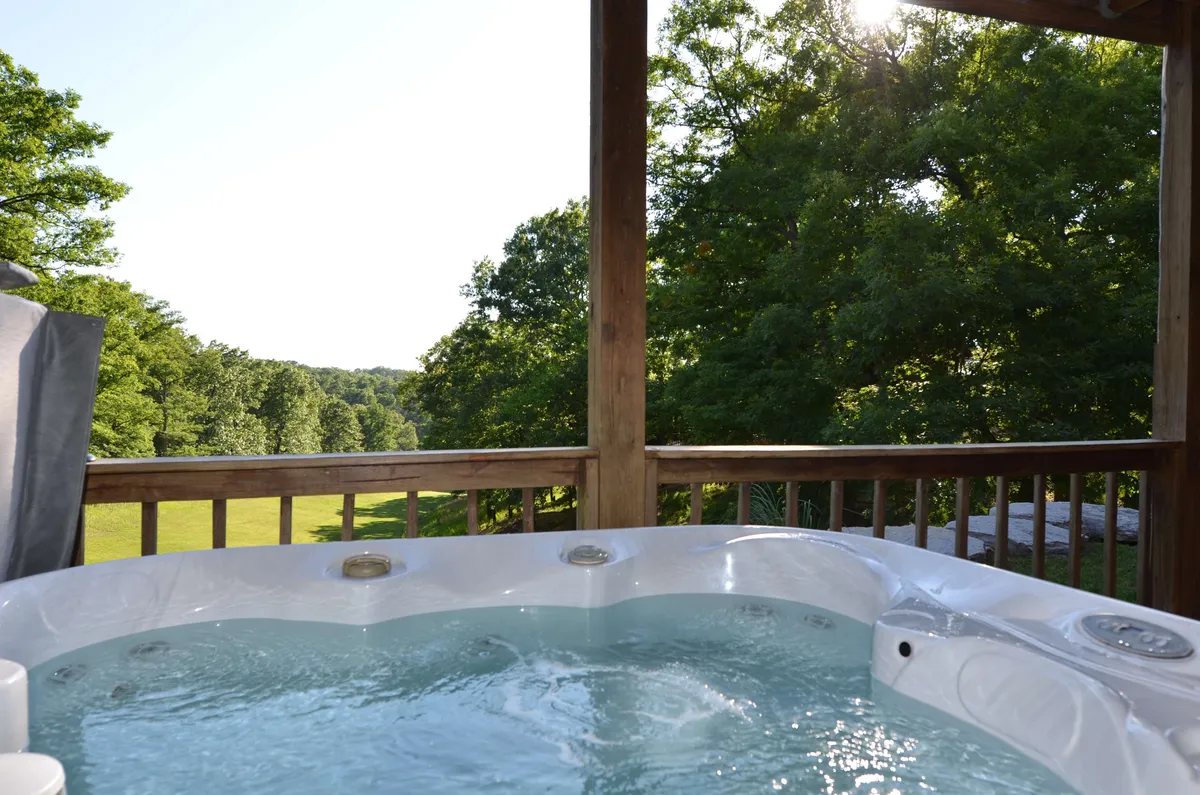 A secluded outdoor hot tub sits on a private deck, surrounded by lush greenery and distant hills. The area feels tranquil, with the hot tub inviting relaxation against the scenic backdrop of the Ozarks.