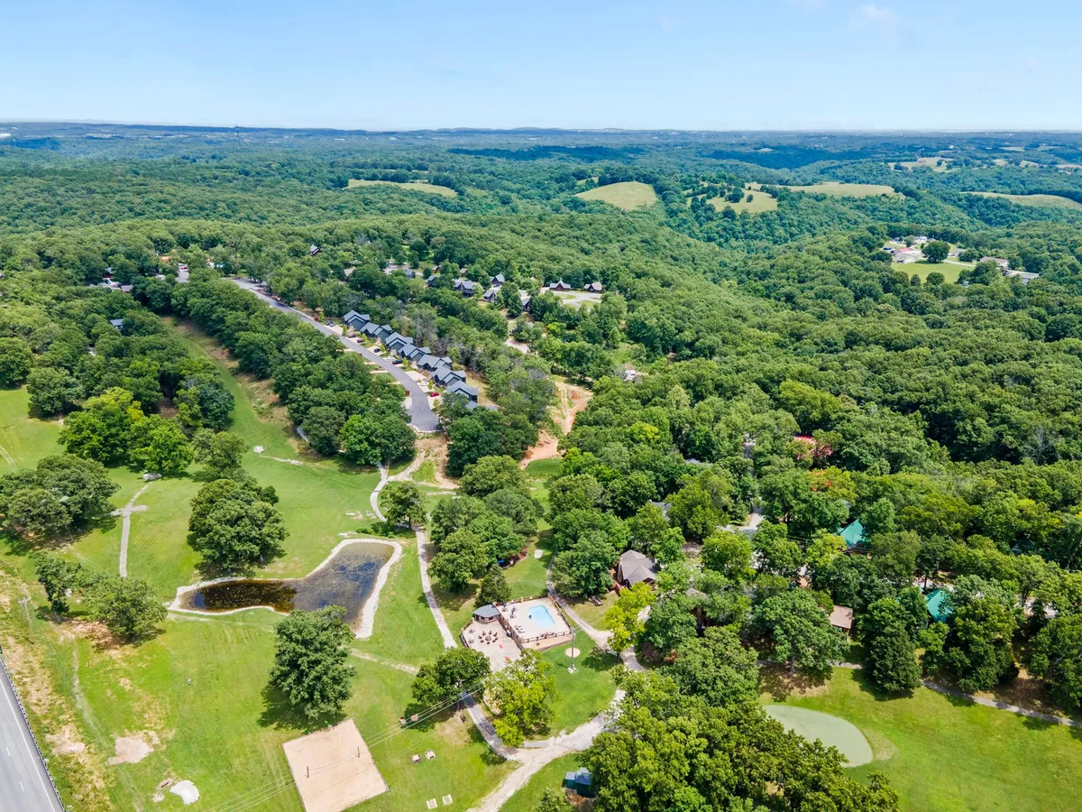 The image showcases a large pool surrounded by lush green trees, a small pond, and rows of cabins in the background. The scene conveys a relaxing resort environment with nature trails and opportunities for swimming or hiking nearby.