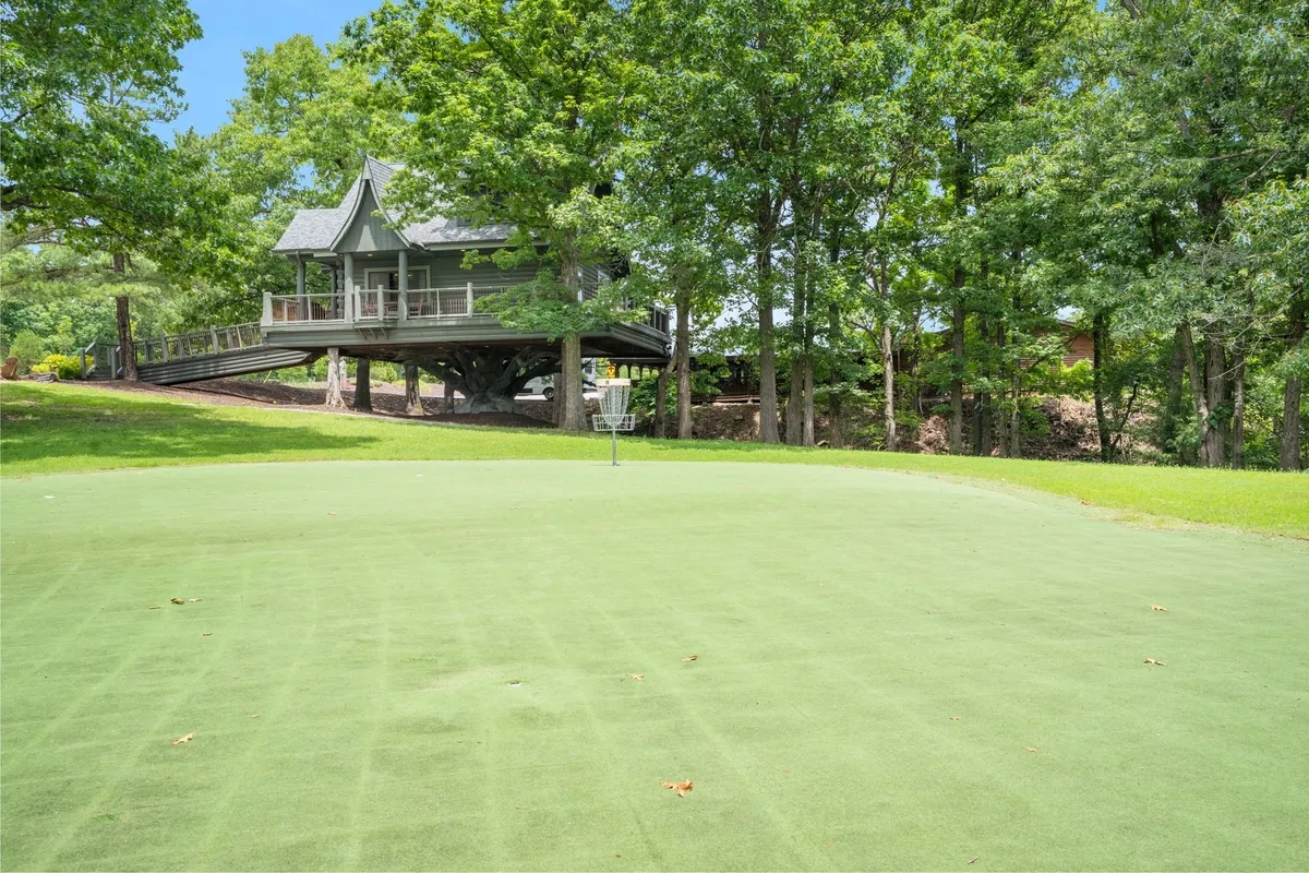 This image features a house on stilts surrounded by lush green grass and trees, with a disc golf basket visible in the foreground. The outdoor setting is peaceful and hints at recreational opportunities right on the property.