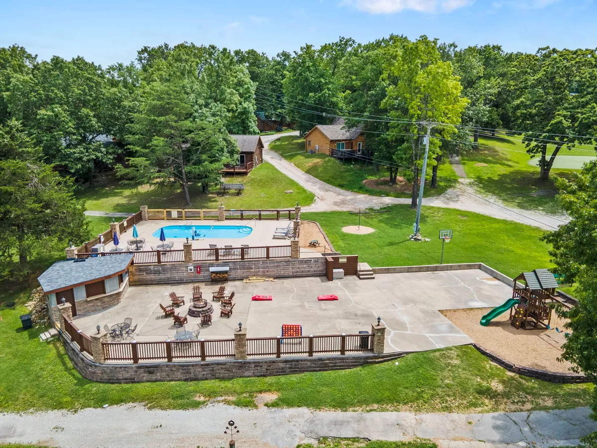 A lively resort space where a sparkling blue swimming pool is bordered by lounge chairs, a playground, and a basketball hoop, all set against a backdrop of green trees. The area is bustling with families enjoying swimming, sunbathing, and outdoor games.