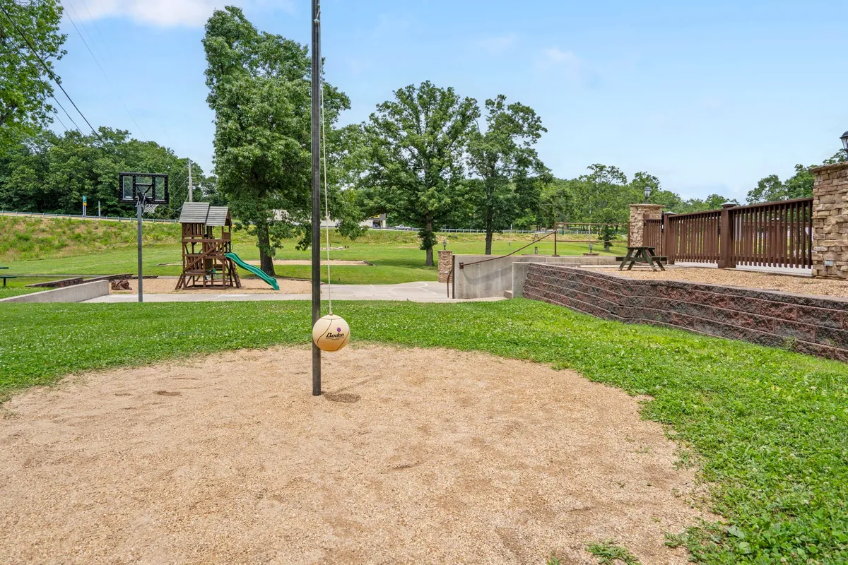 A wide exterior shot captures a communal recreation area featuring a volleyball pole, basketball hoop, playground structure, and picnic area, set against a backdrop of greenery. The space is active and welcoming, designed for various group activities.