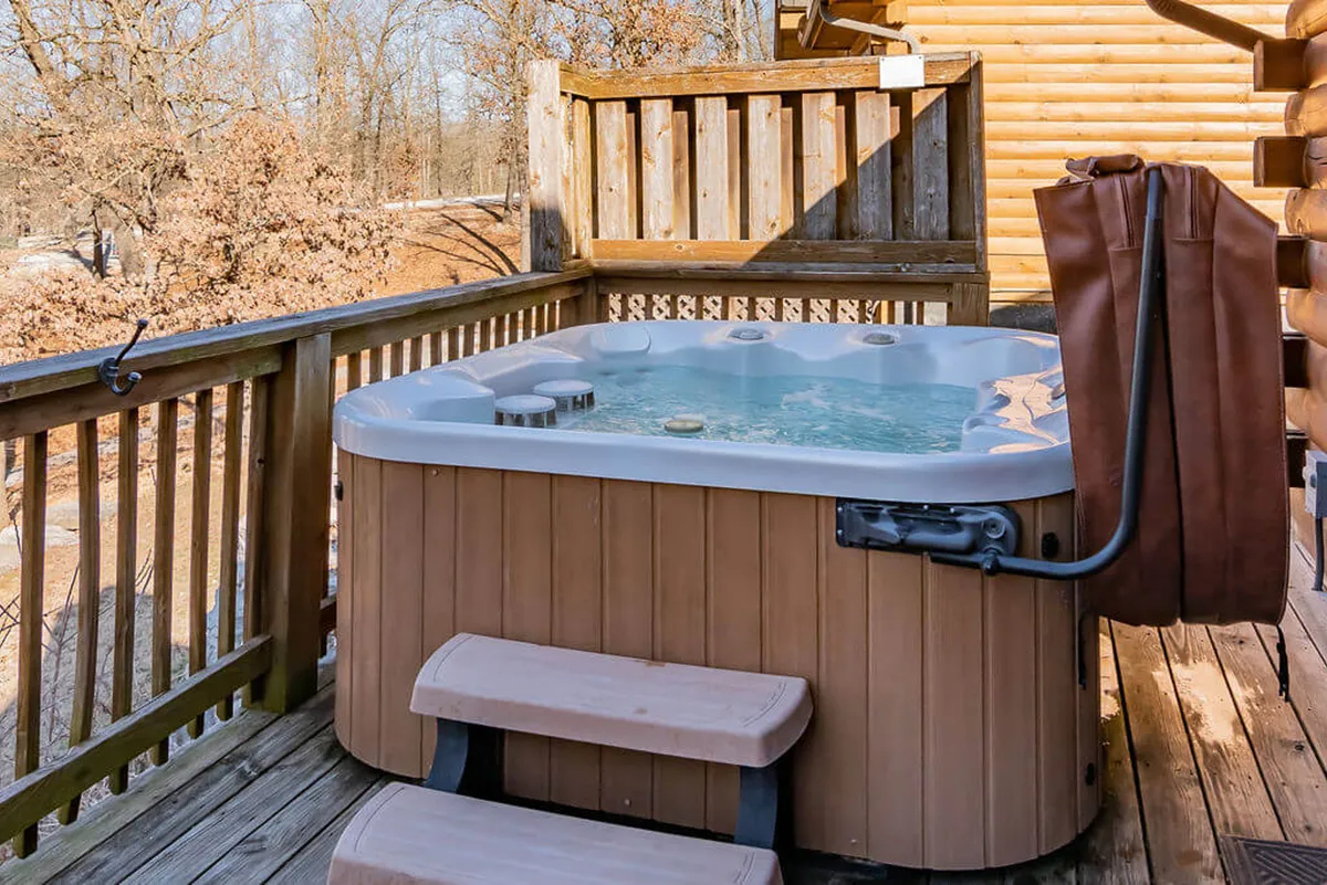 This exterior image captures a hot tub nestled on a wooden deck, surrounded by lush natural scenery. The relaxing setting is perfect for soaking under the stars or watching the sunset over the trees, conveying tranquility and privacy.