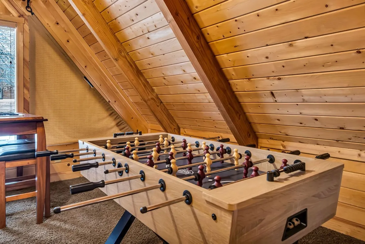 A bright interior loft area showcasing a foosball table beneath rustic wooden beams, a window letting in natural light, and a simple wooden chair nearby. The setting is ideal for friendly competitions and family gatherings, reinforcing the playful spirit of the space.